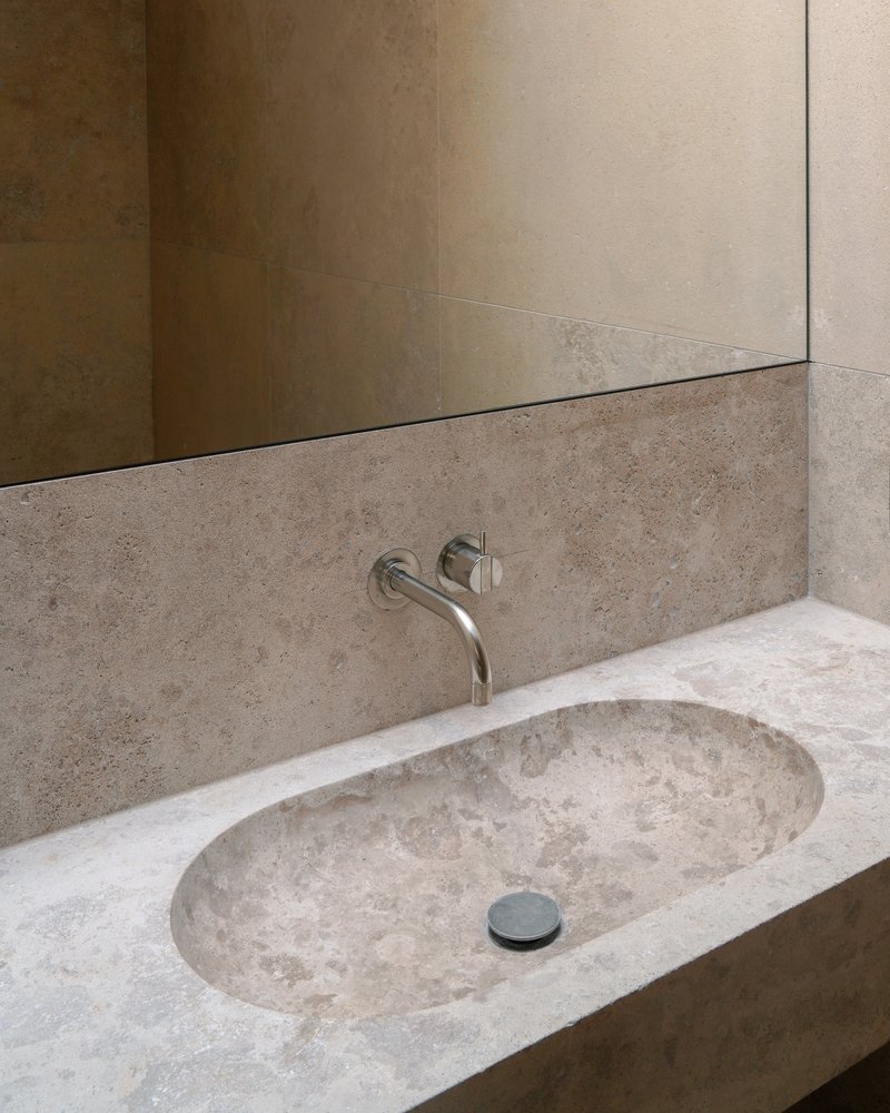 Travertine vanity with integrated oval sink and wall-mounted faucet beneath a frameless mirror