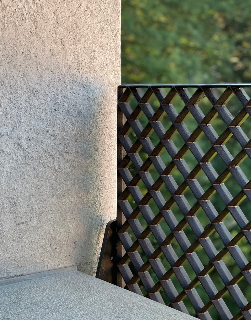 Woven metal screen beside textured plaster wall with blurred foliage beyond