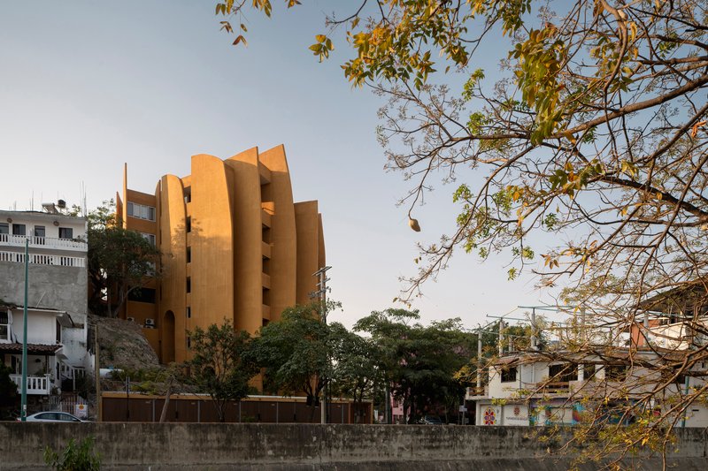 Ochre facade with vertical fin details rising above a stone retaining wall and sparse tree canopy