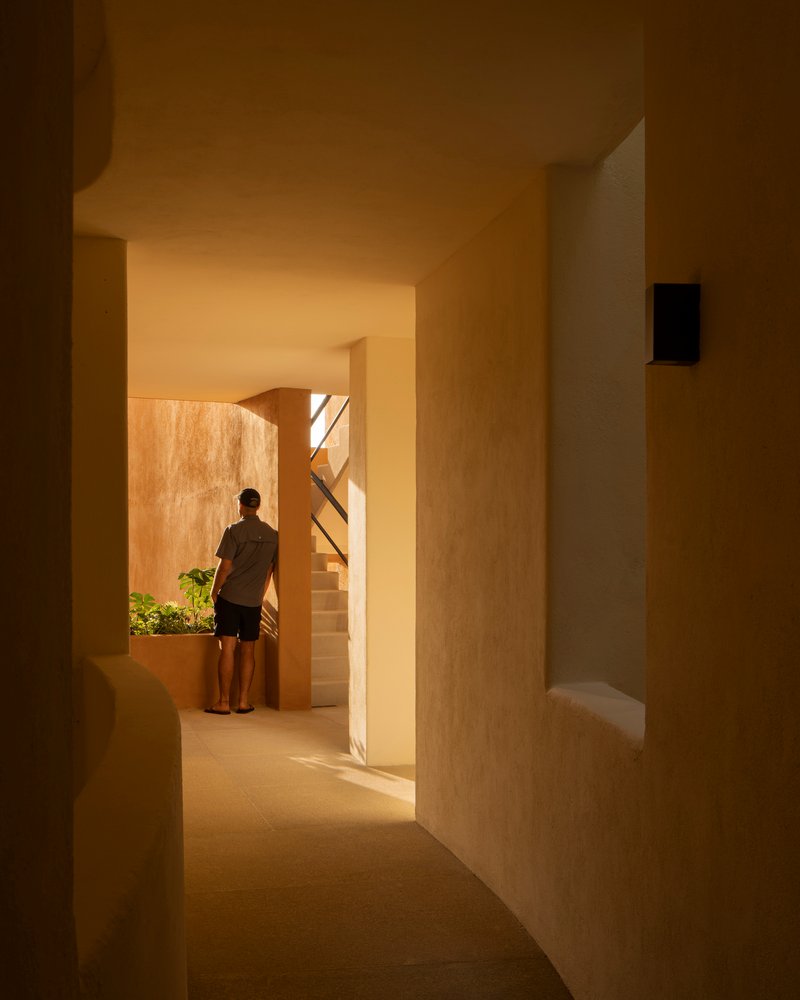 Interior passage with arched openings and warm light revealing a figure standing near a planted courtyard