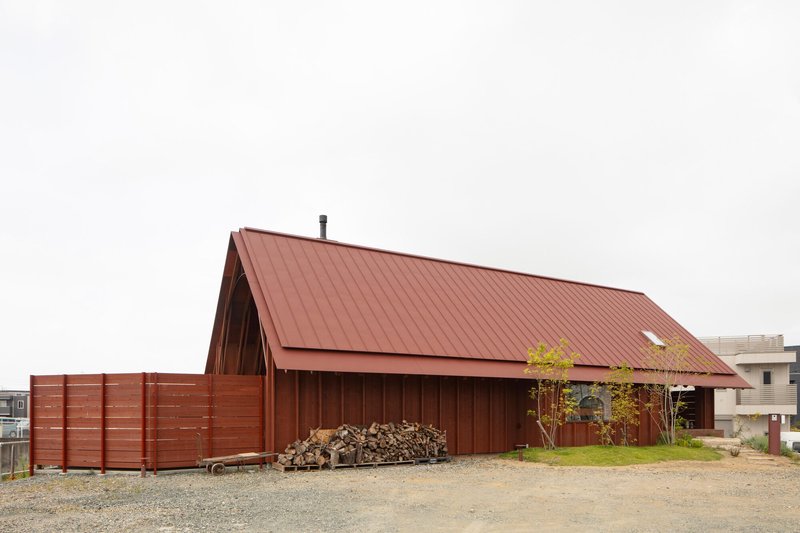 Red corrugated metal gable roof and walls surrounded by a gravel yard with stacked firewood
