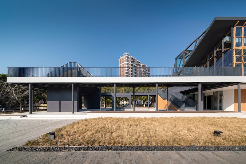 Elevated walkway with vertical screening connecting two volumes above a ground-level plaza in dry grass
