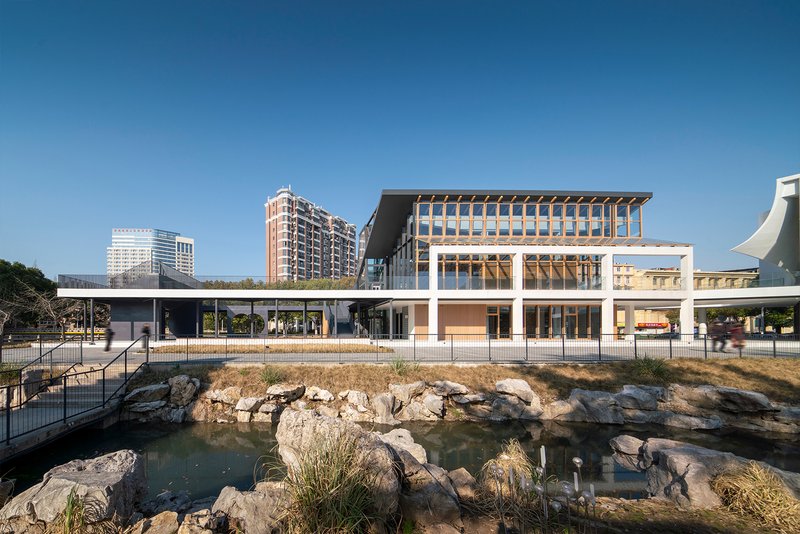 Facade of timber and glass upper volume above colonnade with pond and rocks in foreground