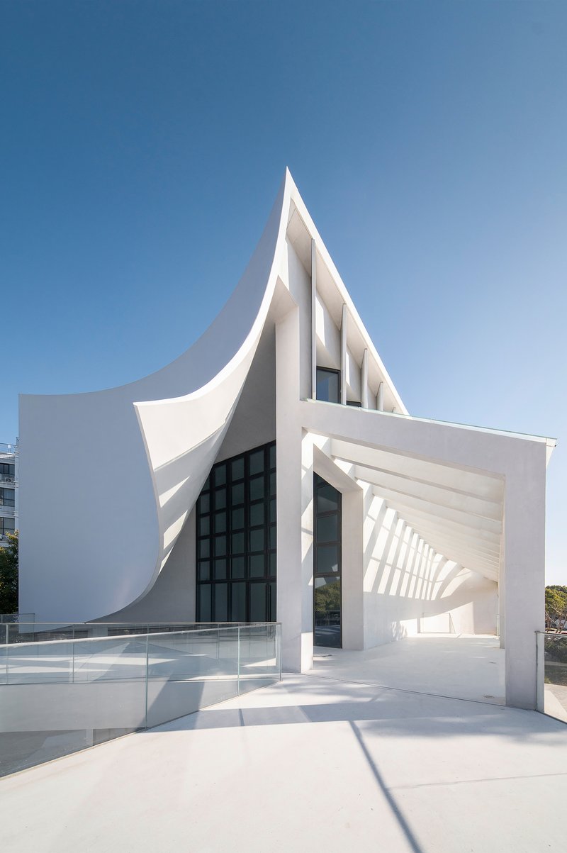 White sculptural entrance with pointed canopy and folded planes framing a glazed opening under clear sky