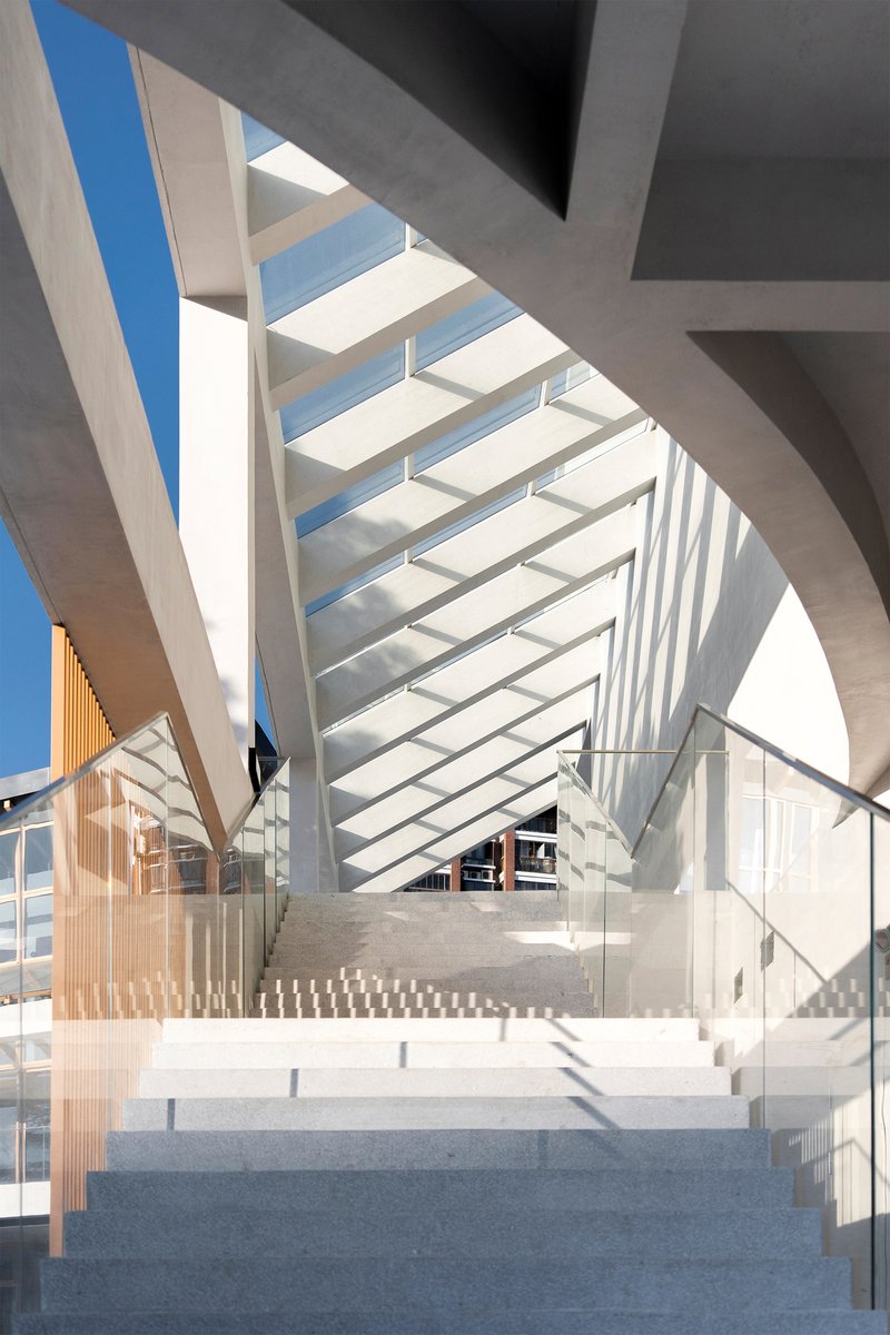 Interior staircase with concrete treads and glass railings beneath a horizontal slatted skylight