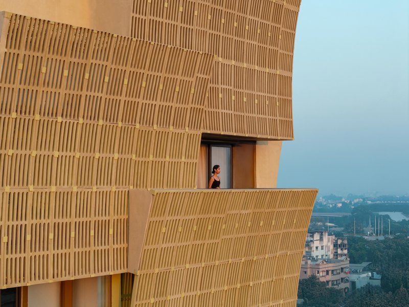 Person standing on a cantilevered balcony framed by ribbed brick screens overlooking the distant cityscape at dusk