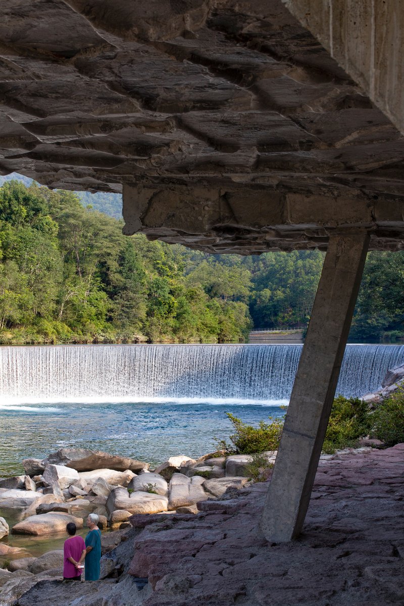 View from beneath stratified rock overhang with concrete column framing a waterfall and two visitors below
