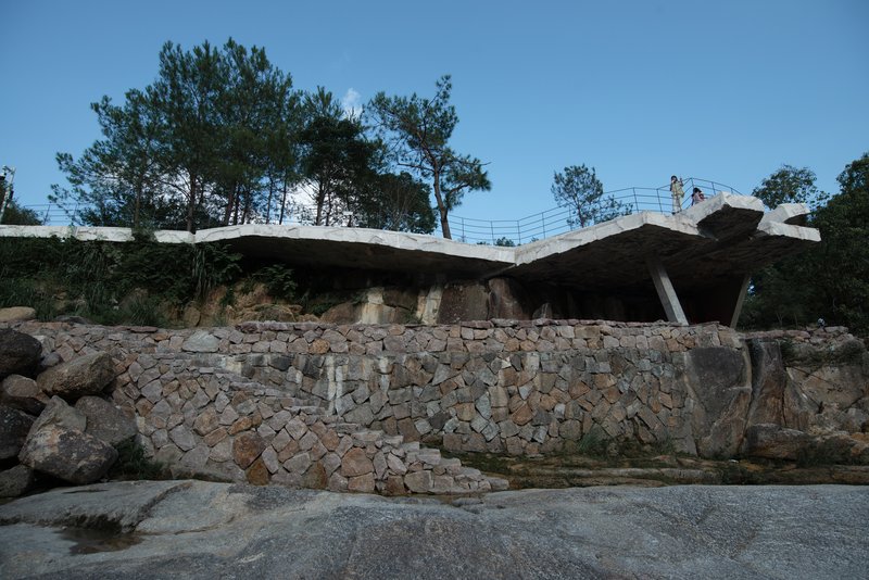 Cantilevered concrete roof slab with exposed underside rising above gabion retaining walls under a clear sky