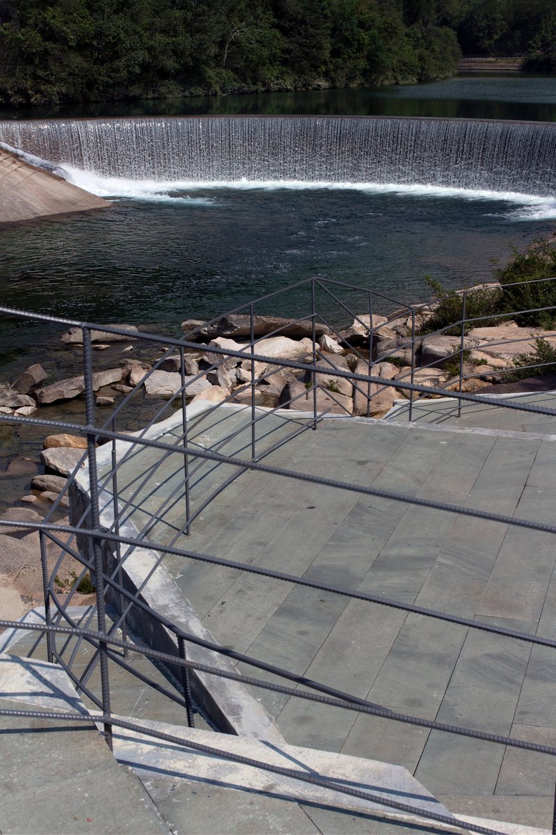 Stone terrace with geometric metal railing overlooking the dam weir and forested hillside