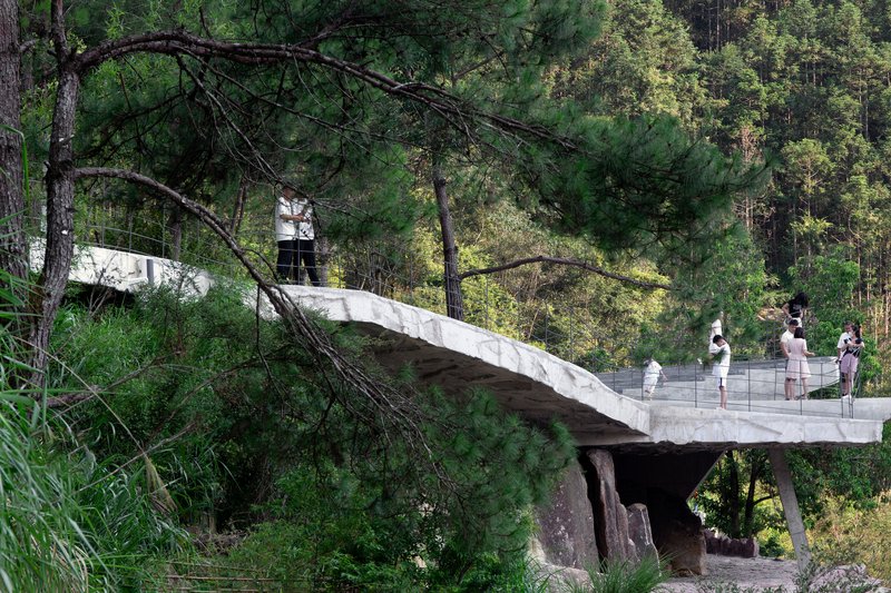 Concrete pedestrian bridge with wire mesh railing extending through pine forest with visitors crossing