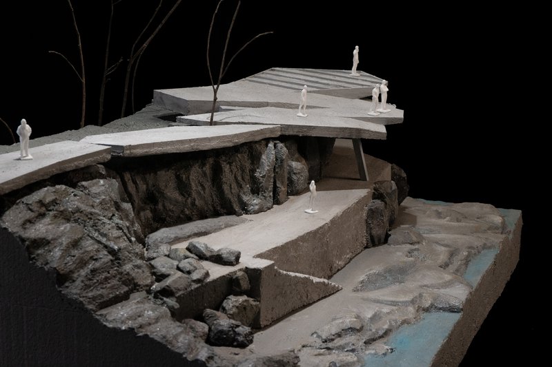 Physical model photographed against black backdrop showing cantilevered terraces over sculpted rocky riverbed with miniature figures