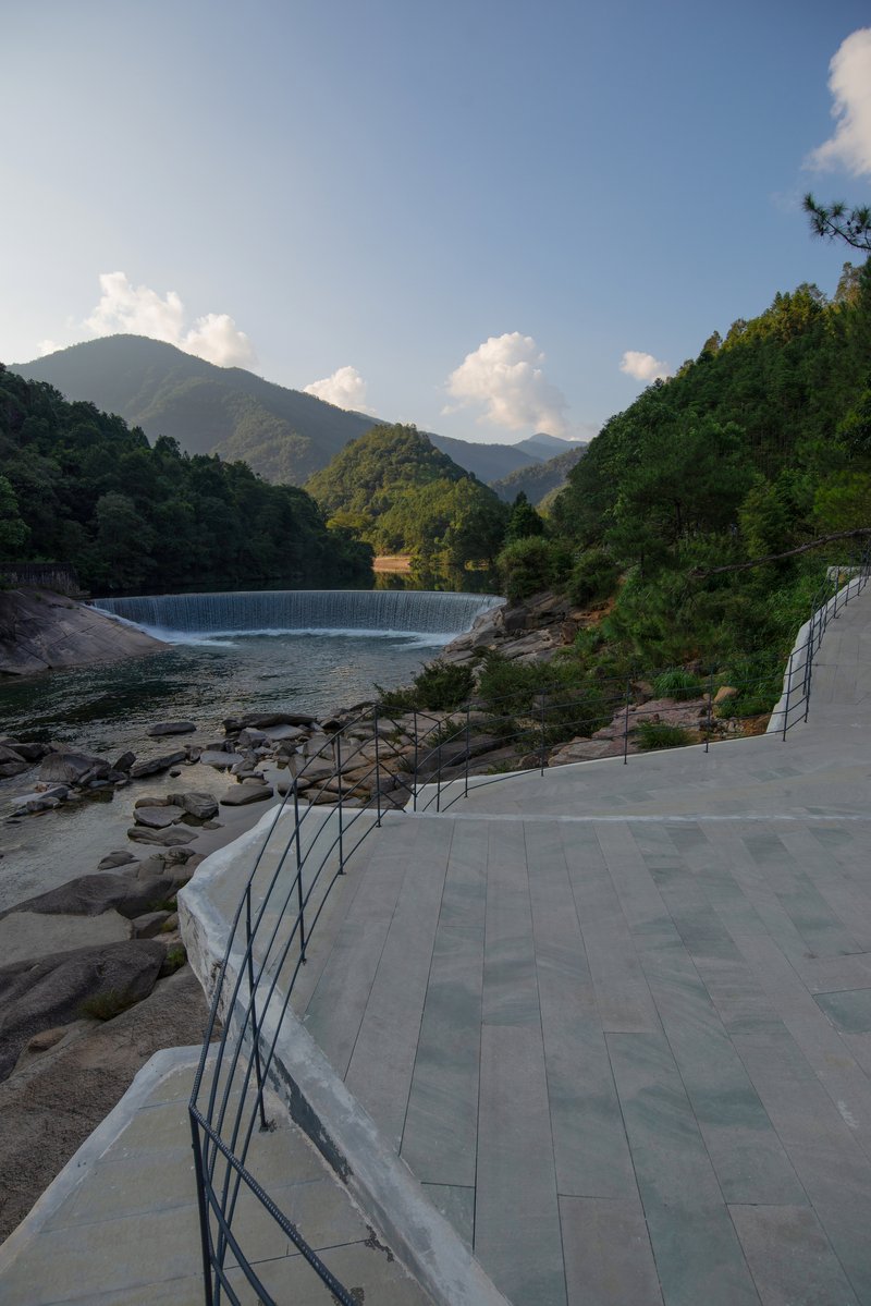 Curving paved terrace with metal handrails following the rocky riverbank below the dam