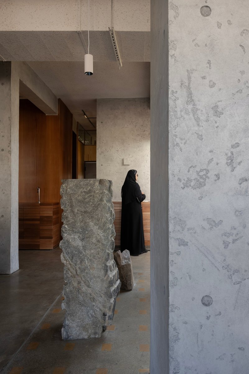 Concrete column and textured wall beside a rough stone fragment with a person standing near timber millwork