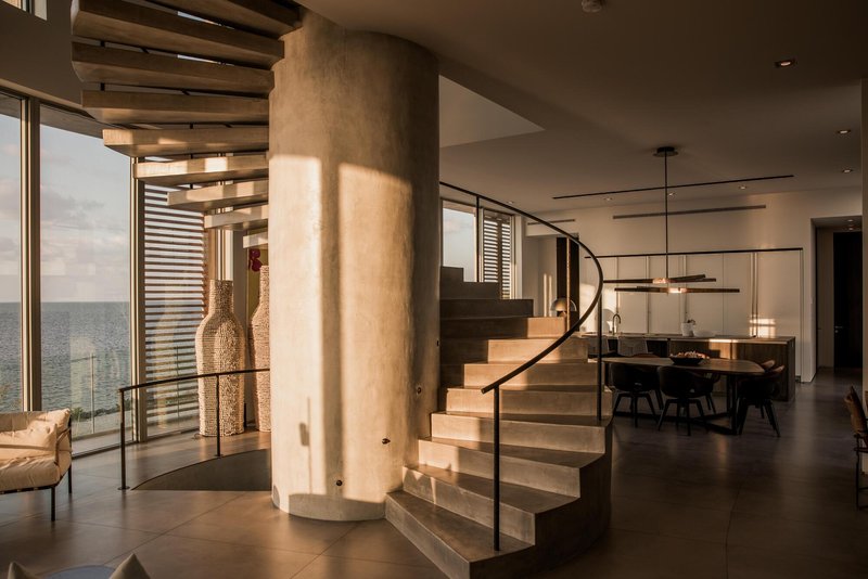 Curved concrete staircase wrapping a cylindrical column with dining area visible beyond at sunset