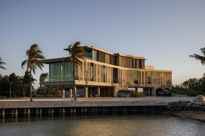 Waterfront elevation with timber and glass volumes elevated on concrete piers under swaying palm trees