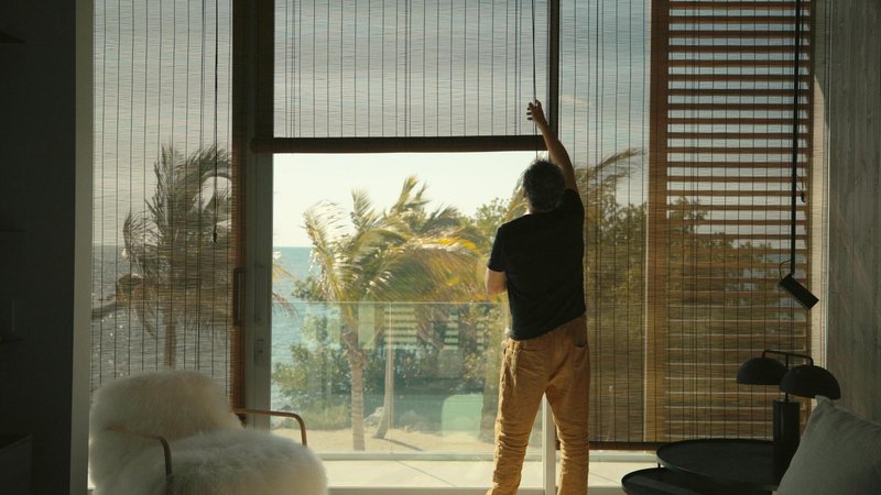Interior view of a person adjusting bamboo blinds at floor-to-ceiling glazing overlooking palm trees and ocean