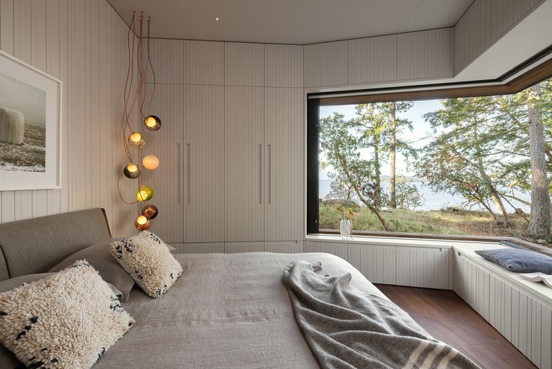 Bedroom with ribbed wall paneling and pendant lights beside a corner window framing coastal trees