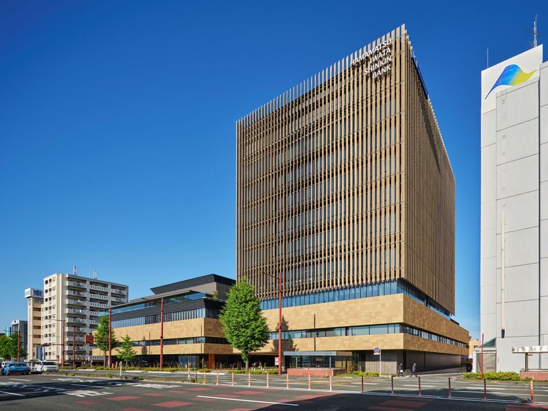 Street view of the vertical timber brise-soleil facade rising above a horizontal stone base under a clear blue sky