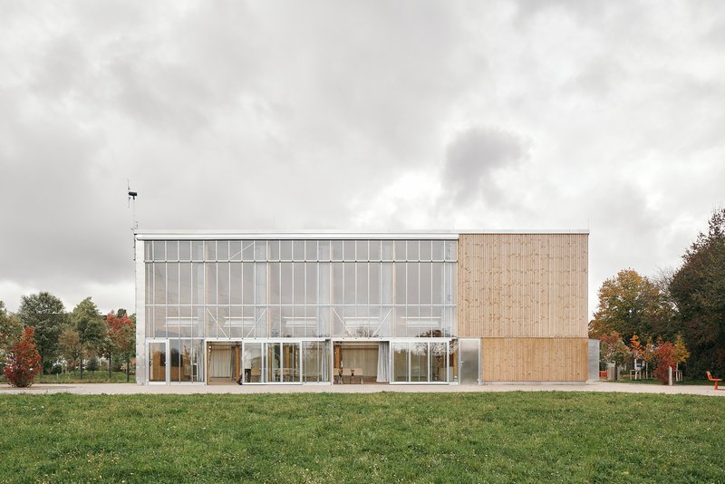 Long west elevation with the full-height greenhouse facade alongside a closed timber block