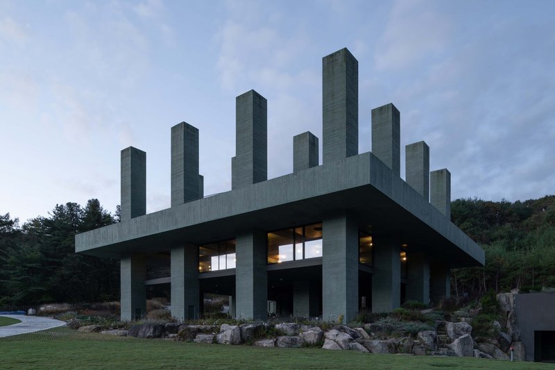 Green concrete structure with vertical towers illuminated from within at dusk surrounded by boulders and grasses