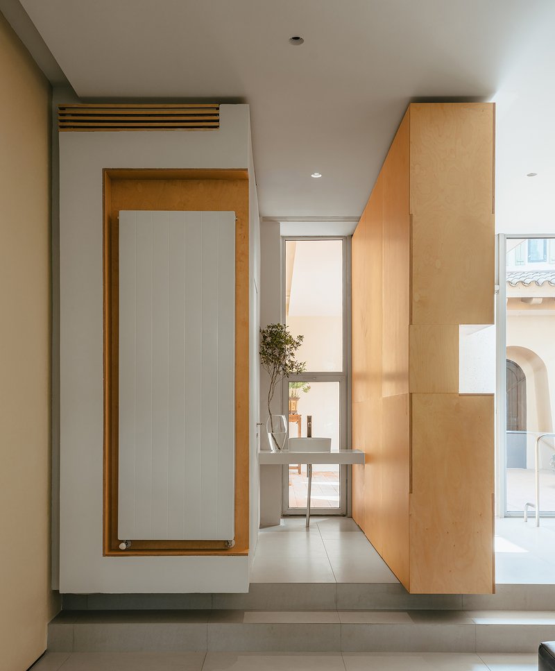 Hallway with plywood radiator panel and glass door overlooking courtyard in afternoon light