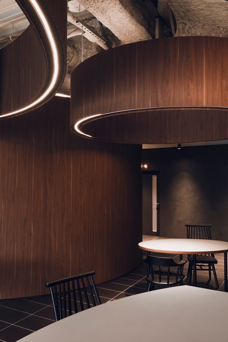 Dining area with circular walnut veneer volumes and illuminated ring details suspended from an exposed concrete ceiling