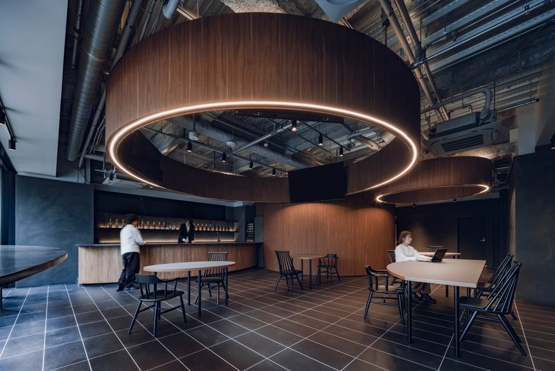 Dining area with curved wooden pods and illuminated ring fixtures as two figures move through the space