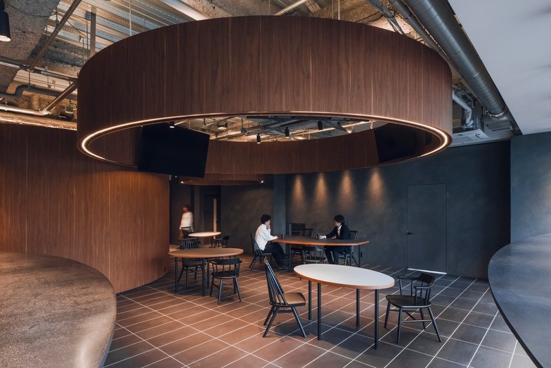 Lounge area where dark tile meets timber flooring as two people sit at tables beneath ring lighting