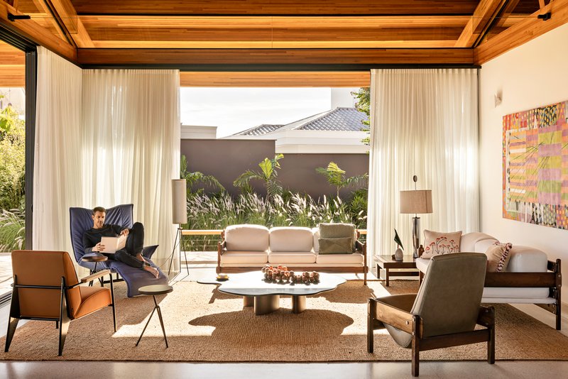 Living room framed by floor-to-ceiling curtains opening to a planted courtyard with a seated figure reading
