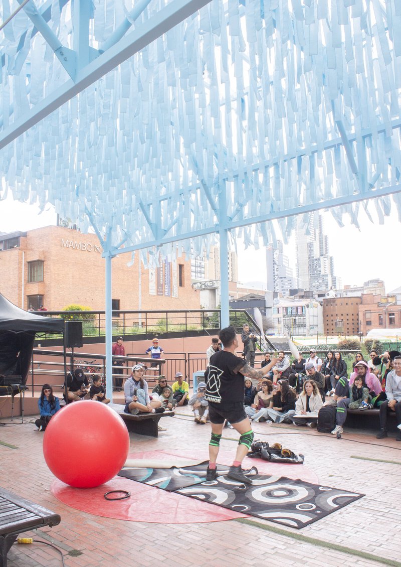 Translucent blue canopy overhead sheltering a public performance space with seated audience and large red ball