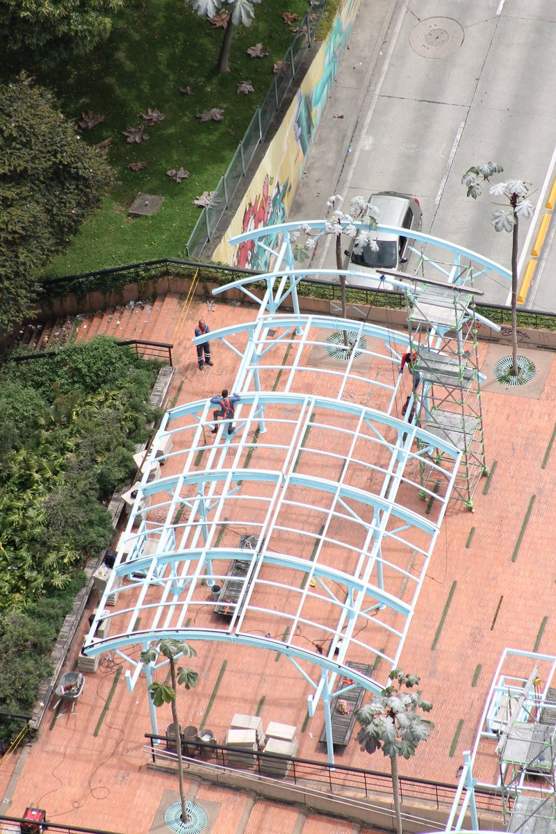 Overhead view of one white steel canopy frame arching over terracotta paving with people and planted pots