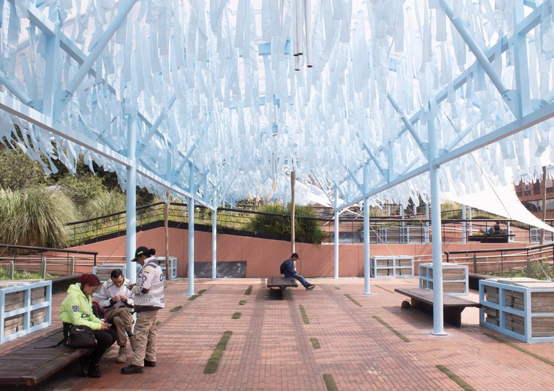 Covered plaza with tiled paving and benches beneath a canopy of hanging blue panels