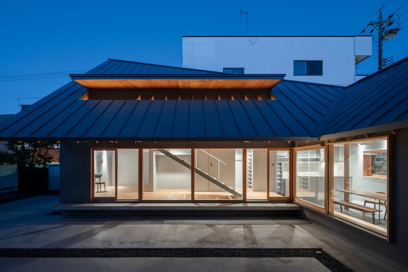 Evening view of the glazed facade beneath the metal roof with interior lighting and visible staircase