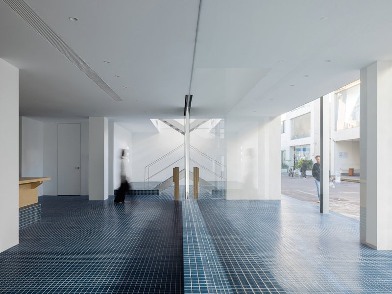 Double-height foyer with blue mosaic tile floor, timber columns, and figure passing through sunlit space