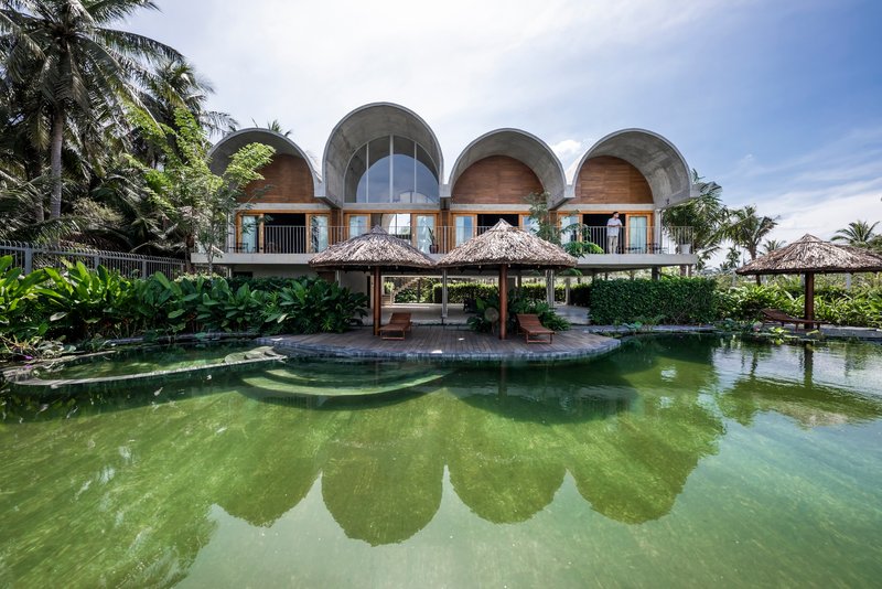 Arched concrete facade with thatched pavilions reflected in a green pool surrounded by palms