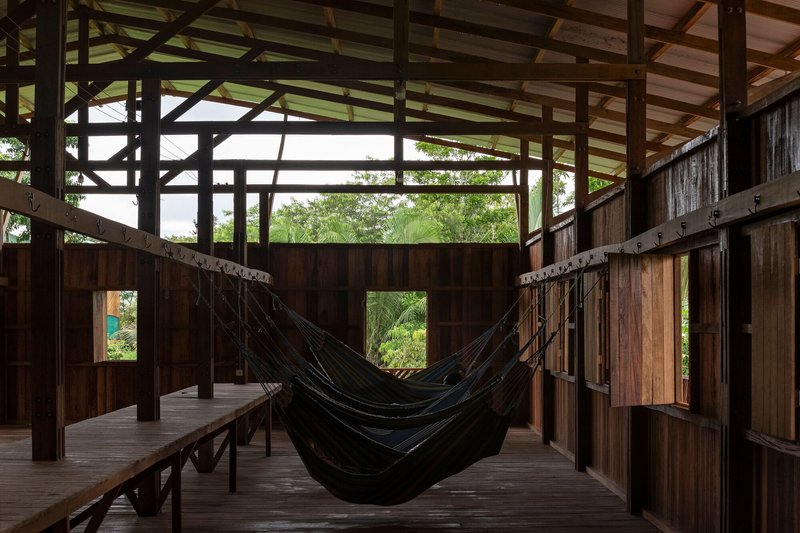 Interior space with suspended hammock between timber walls and exposed structural frame opening to vegetation