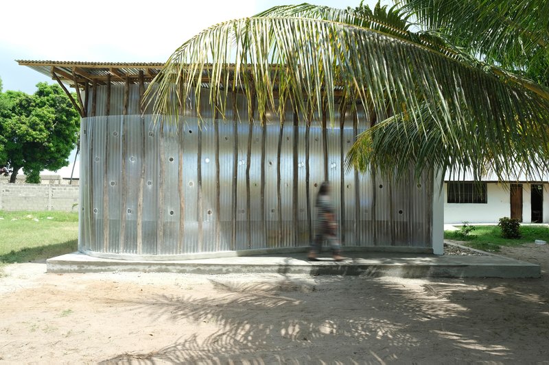Curved corrugated metal facade with circular openings beneath overhanging palm fronds and dappled shade