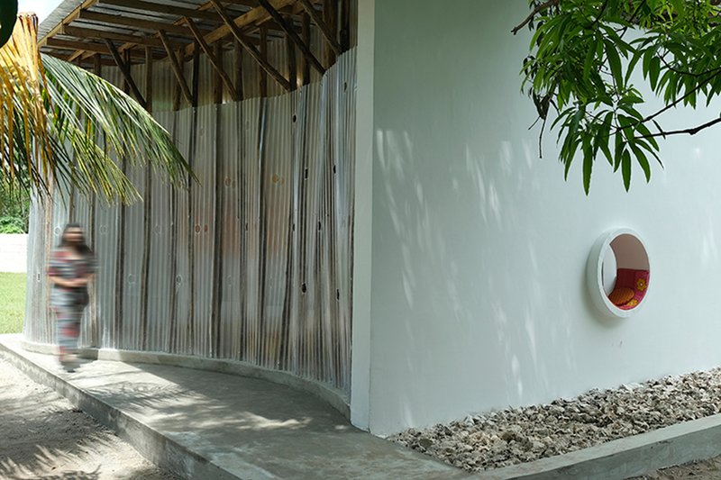 Covered walkway with vertical timber screens and a circular window opening as a figure passes through dappled light