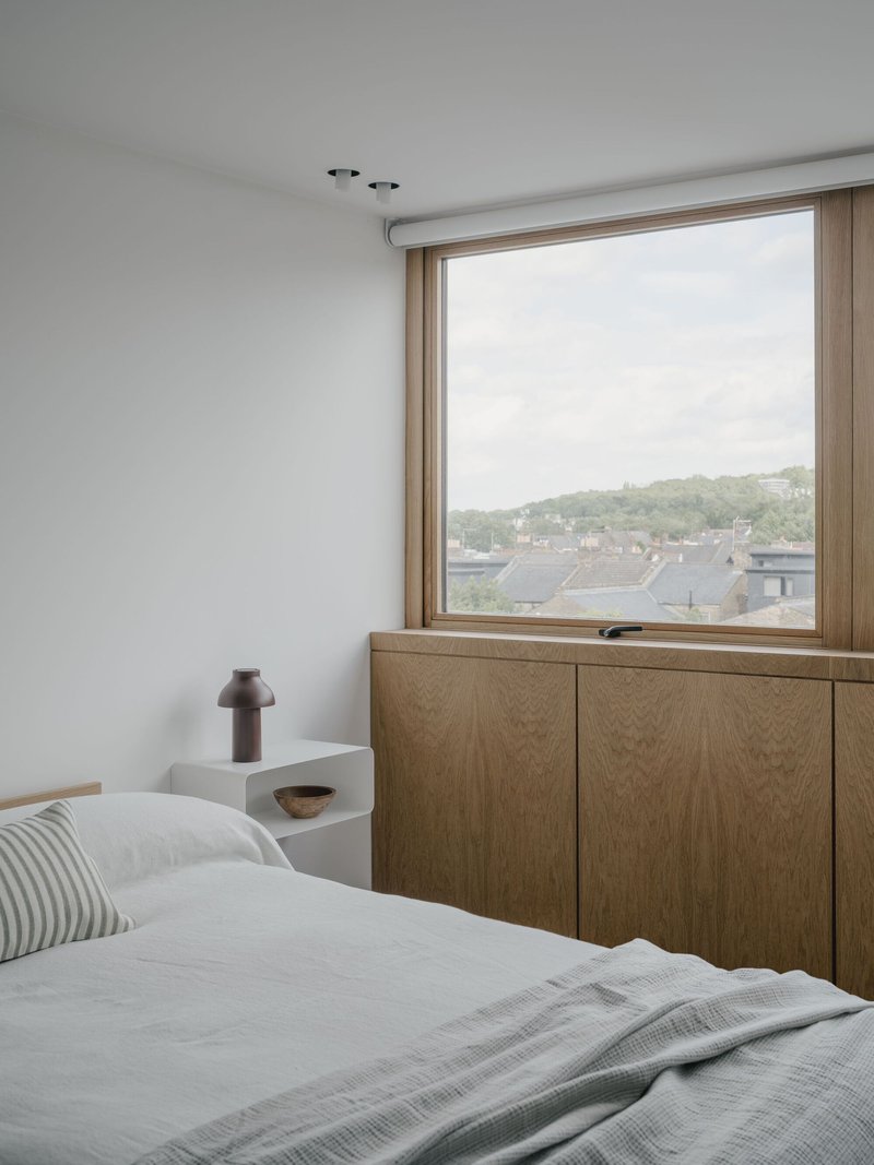 Bedroom with oak cabinetry below a large timber-framed window overlooking rooftops and trees
