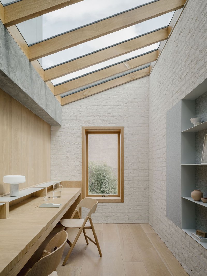 Study with timber beam skylight strips above white brick walls and vertical window overlooking greenery