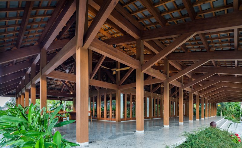 Open-sided pavilion with exposed timber trusses and columns framed by tropical plantings