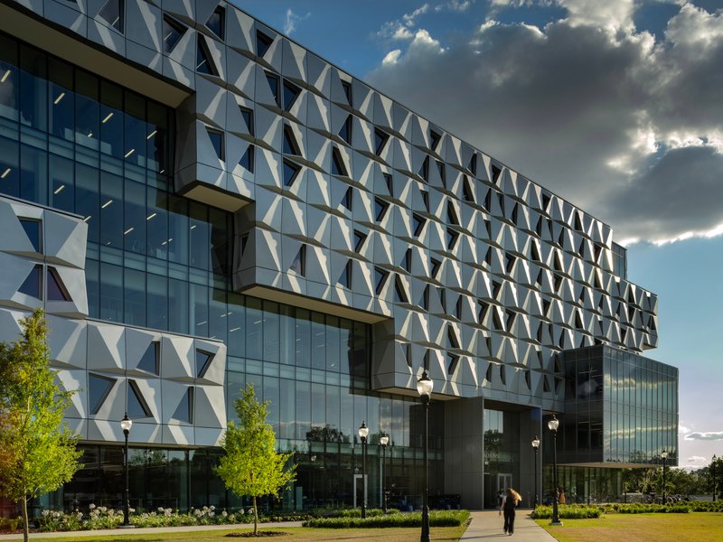 Glass and white triangular-paneled facade under dramatic clouds with pedestrians on the lawn below