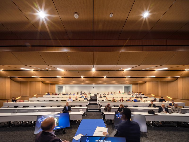 Tiered lecture hall with undulating wood ceiling panels and recessed strip lights illuminating rows of students