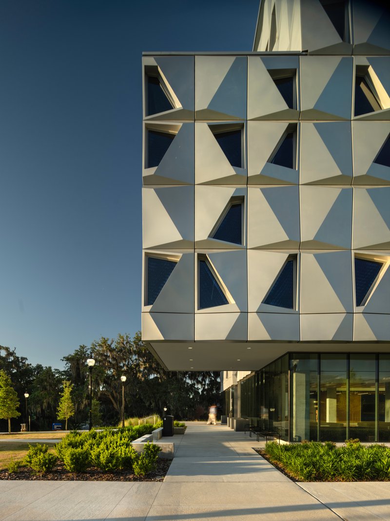 Geometric folded panel facade above glass-walled ground floor and landscaped entry plaza at dusk
