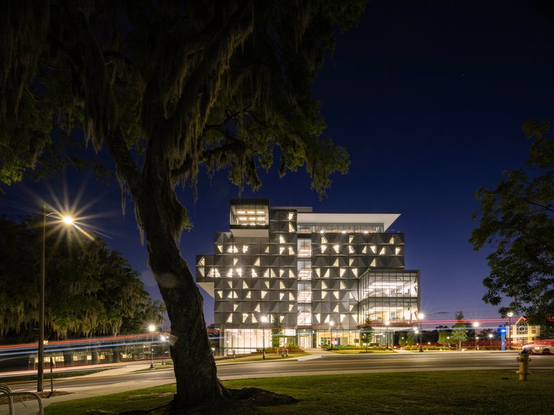 Multi-story glass and perforated metal facade illuminated at dusk beside a moss-draped oak tree