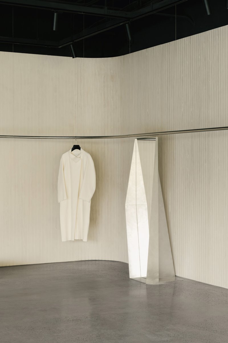 Interior corner with pale ribbed timber walls, concrete floor, and garments hanging near a white angular partition
