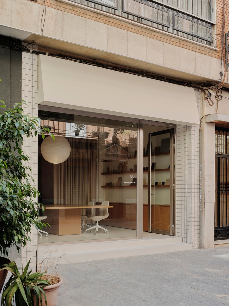 Street-facing storefront with white tile surround, retractable awning, and glazed opening revealing office interior with potted plants