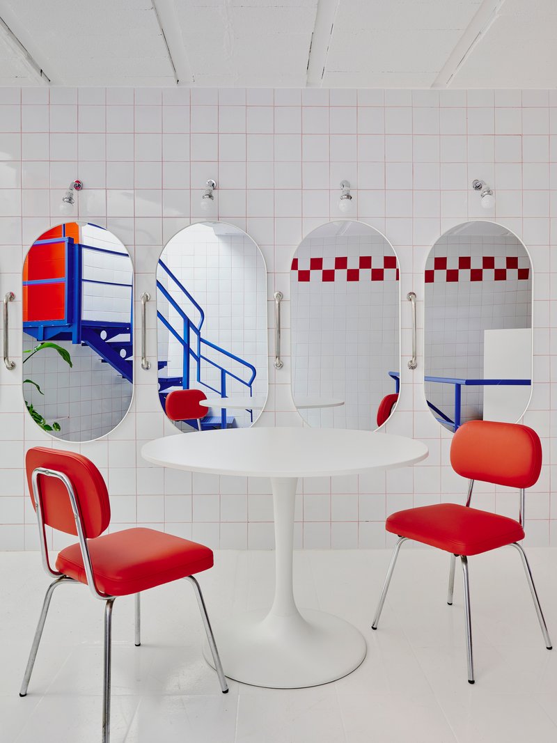 White tiled room with four oval mirrors bearing graphic patterns, white pedestal table, and red upholstered chairs