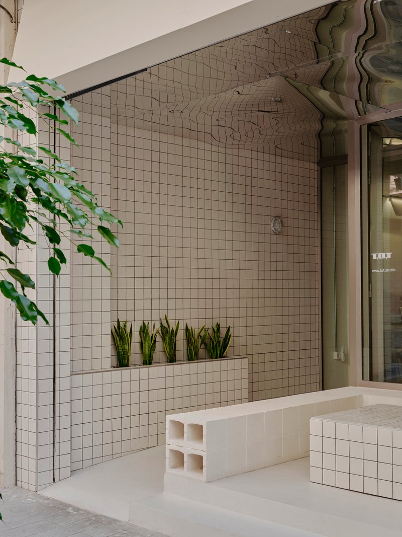 White grid-tiled courtyard wall with integrated planter shelf, trailing vines overhead, and modular tile seating blocks