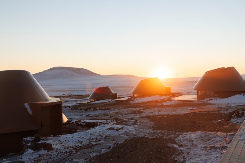 Cluster of dome-roofed structures on snow-covered volcanic terrain at sunrise with mountains in the distance